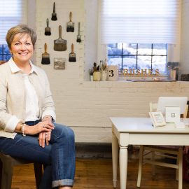 Donna Thomson, photographed by Portland Headshot, standing among some of the hand painted furniture that she creates and sells at the Pepperell Mill Campus.